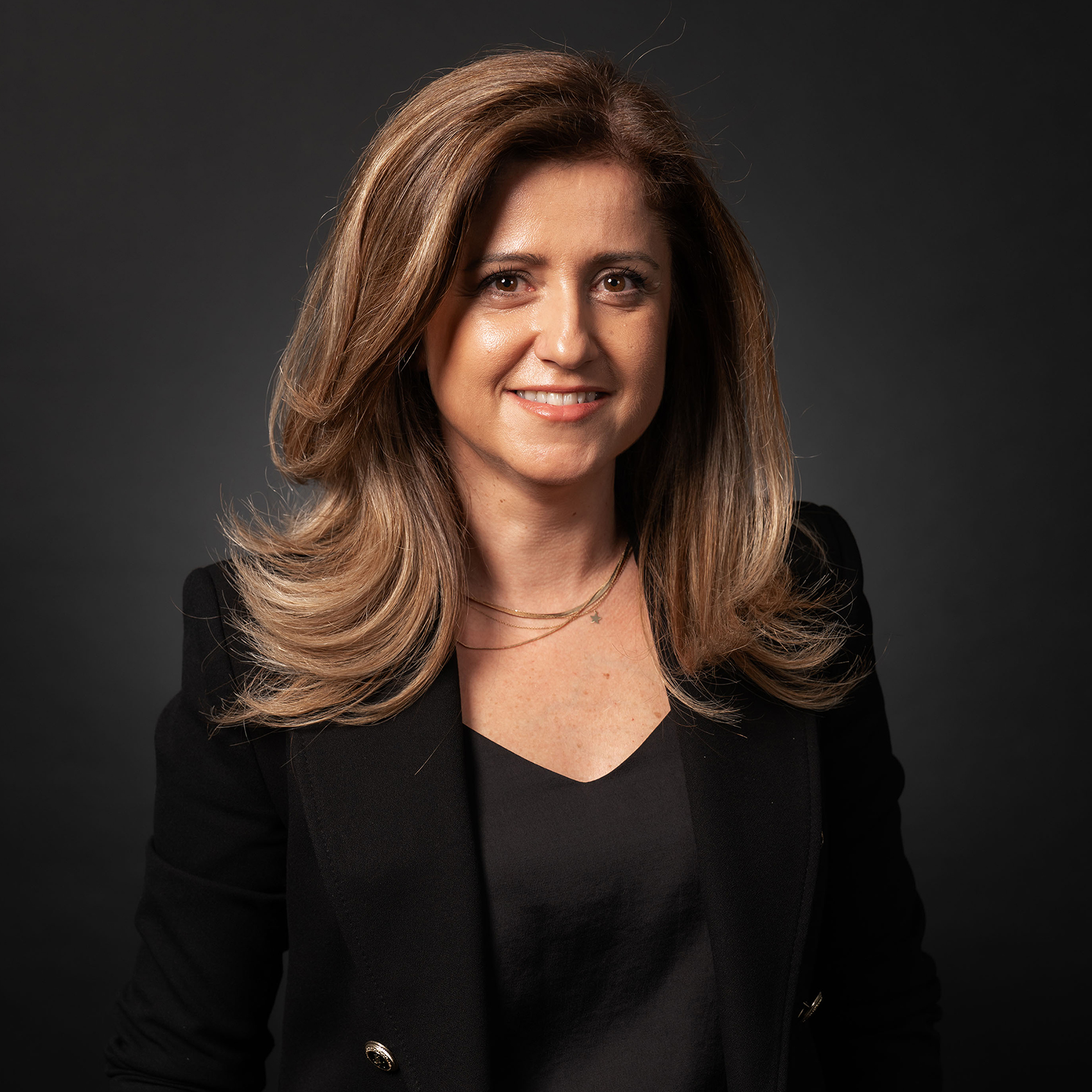 A smiling professional woman in a bright red/orange jacket, with long dark curly hair.